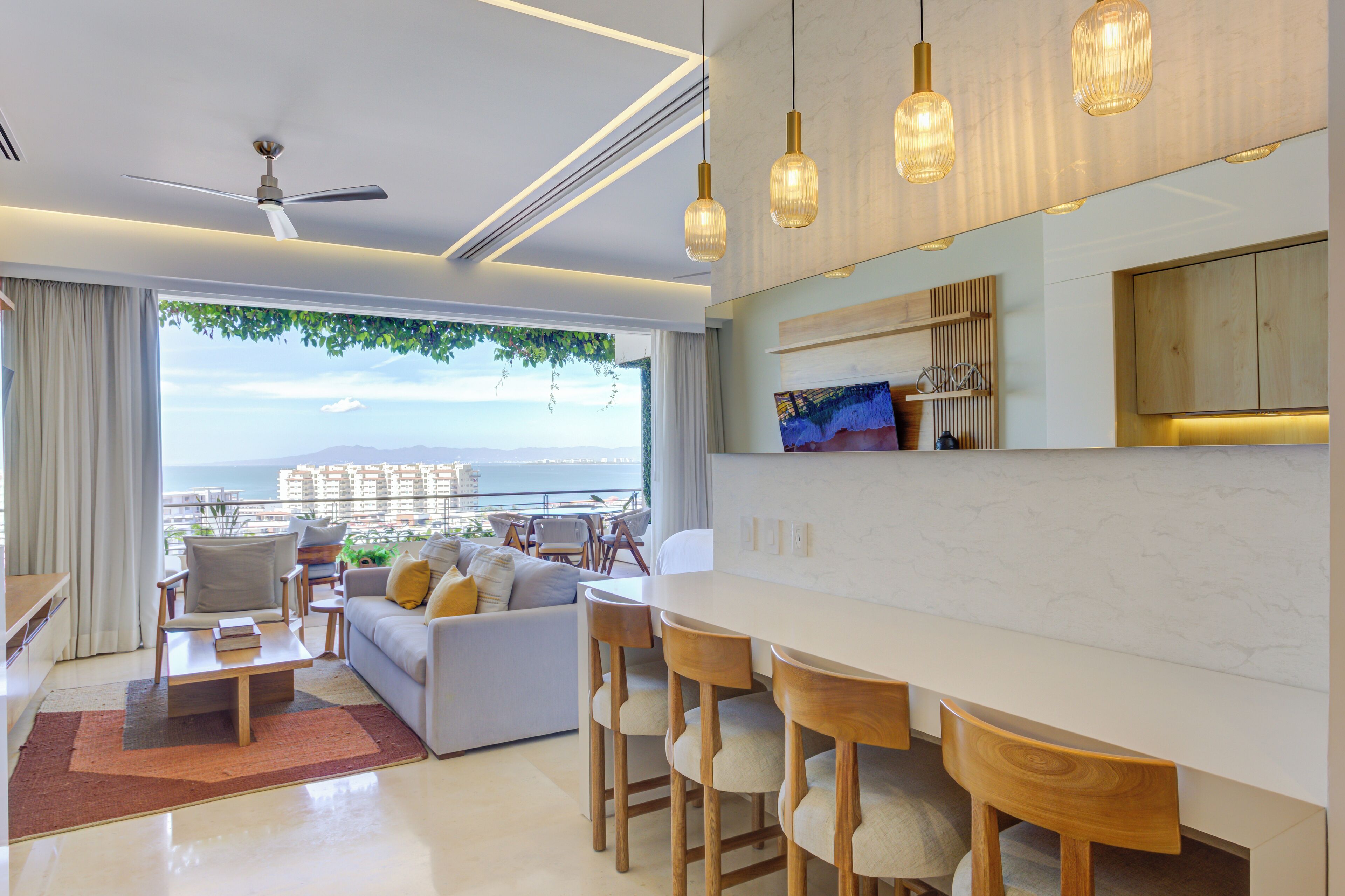 Un soggiorno moderno con divano, poltrone, tavolino da caffè e zona pranzo con sgabelli da bar. Sono presenti una ventola a soffitto, lampade a sospensione e vista sull'oceano attraverso ampie finestre.