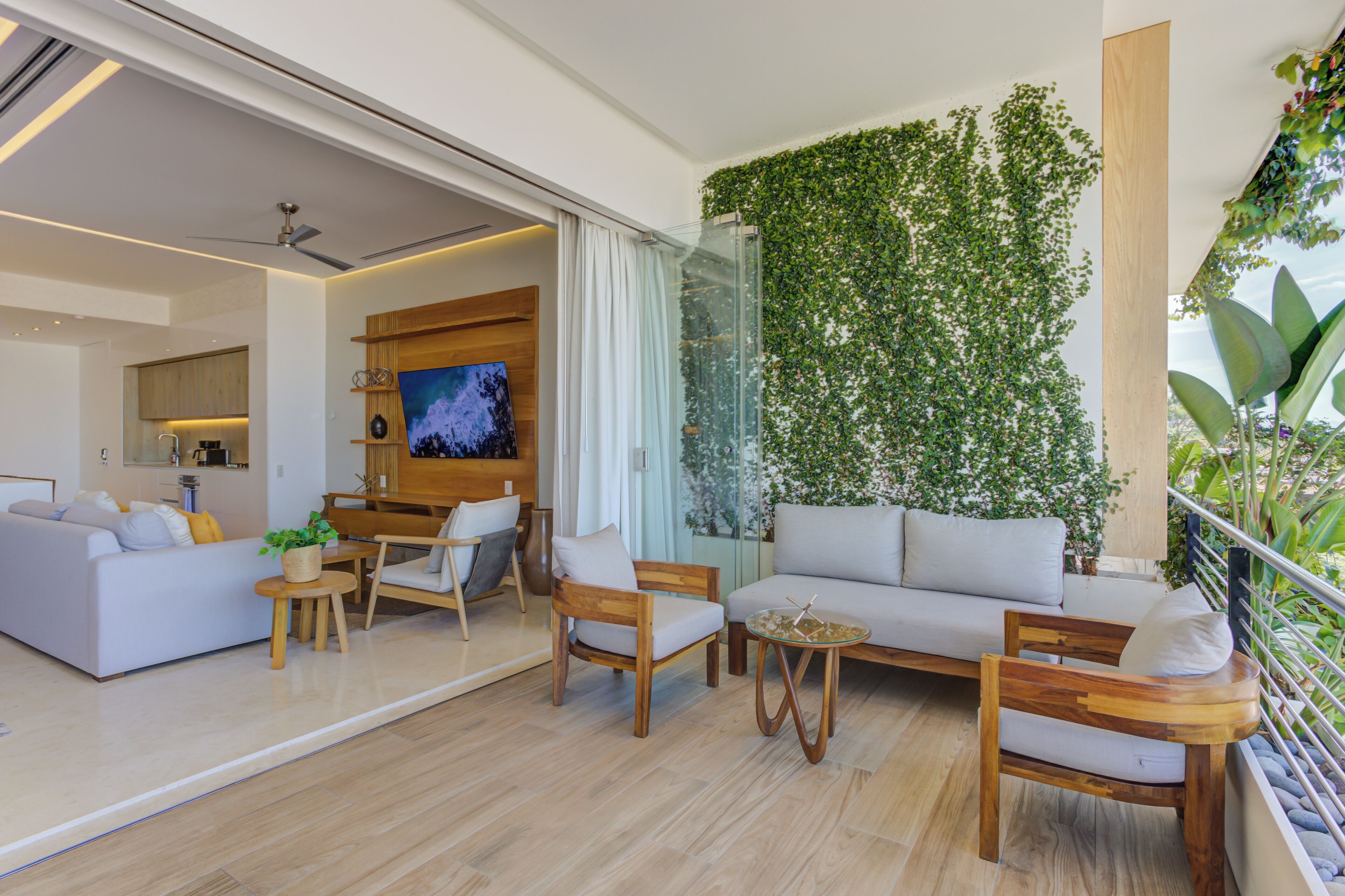 Una sala moderna con sofá, sillones, una mesa de centro y televisor. Hay una puerta de vidrio que da a una cocina y un balcón con una pared verde.