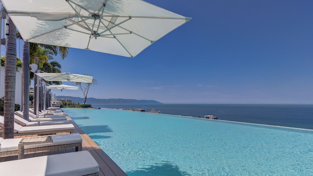 A poolside area with lounge chairs and umbrellas, overlooking a clear blue ocean. - Elegant Condo SohoPV Ocean Views & Rooftop Pool Short Description (Puerto Vallarta)