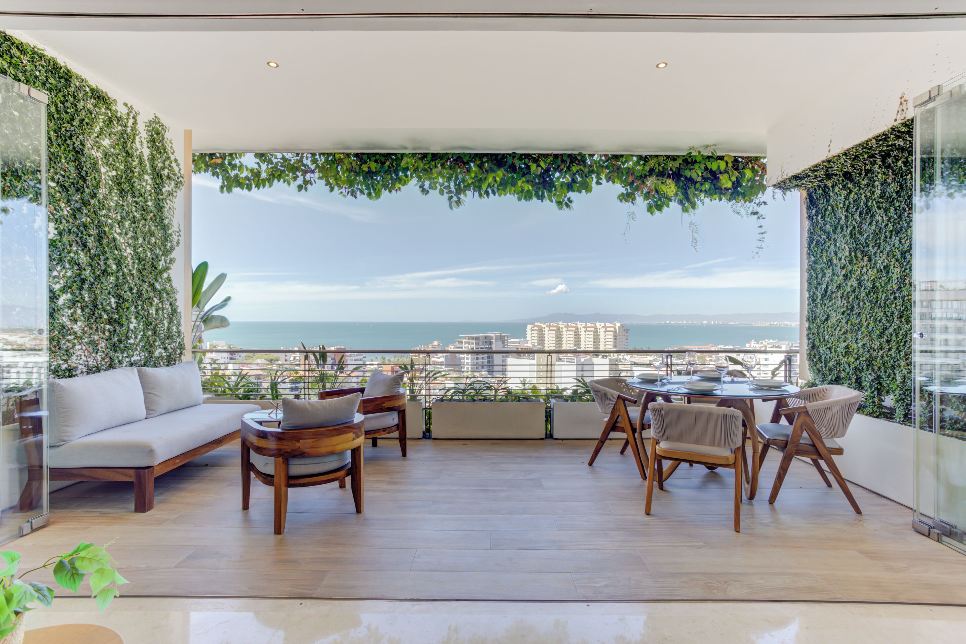Un balcon moderne aménagé avec un canapé, des fauteuils et une table à manger, offrant une vue sur le paysage urbain et la mer.