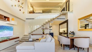 Un vaste espace de vie comprenant un canapé blanc, un téléviseur à écran plat et une table à manger avec des chaises. Un escalier mène à l’étage supérieur.