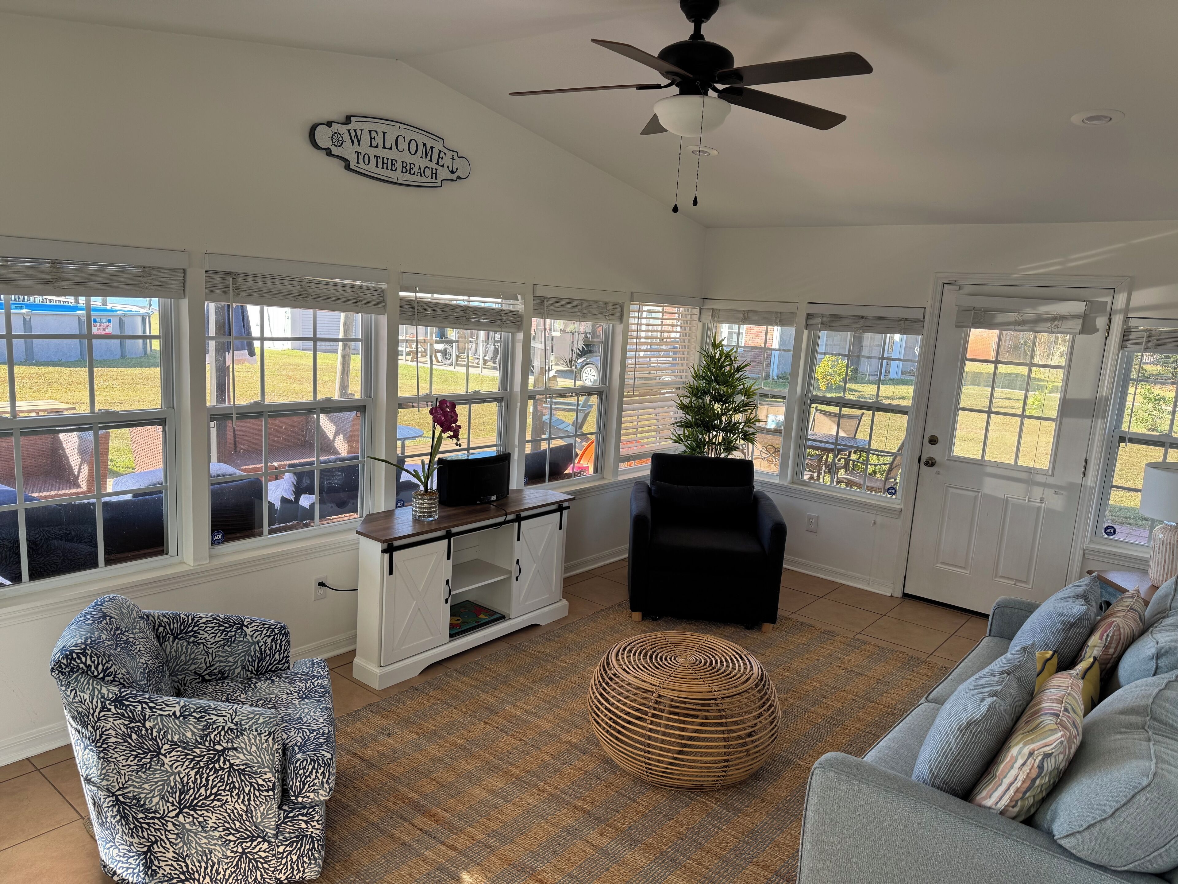 A sunlit room with a view of the backyard, a ceiling fan, a white console table, a black armchair, a wicker ottoman, and a grey sofa with patterned cushions.