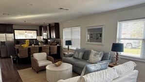 A modern living room with a grey sofa, a white armchair, and a round coffee table. There is a kitchen area with dark wooden cabinets and a stainless steel refrigerator. - Nice House in Perdido Bay (Lillian)