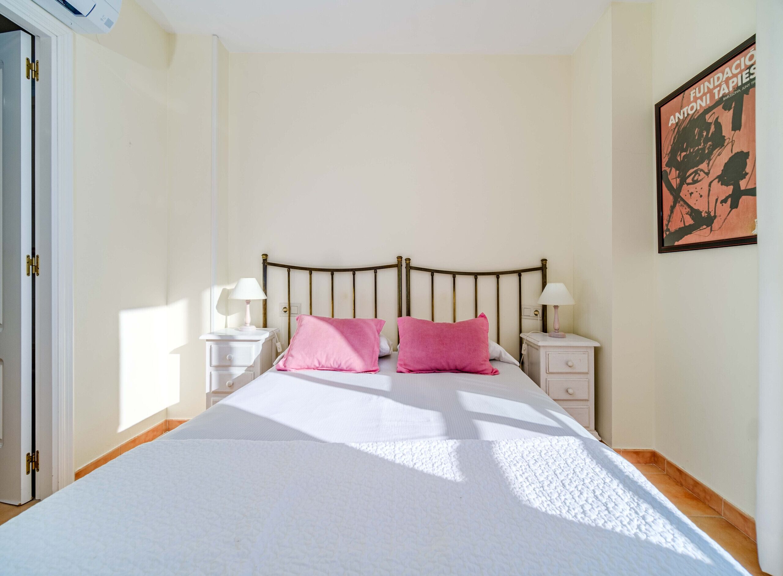A bedroom with a bed, two bedside tables with lamps, a door, and a framed artwork on the wall.