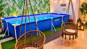 Un bain à remous bleu orné du logo « INVTEX », deux fauteuils en osier, une petite table et un mur décoré d’un motif de feuilles tropicales.