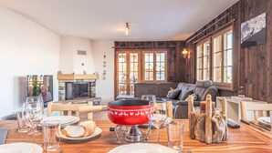 Ein Essbereich mit einem Holztisch, der für eine Mahlzeit gedeckt ist, einem Kamin und einer Wohnbereiches mit Sofa und Fenstern.