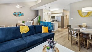 A modern living room with a blue sofa, yellow throw, and a dining area with wooden chairs and a table. - Resort Pool, 5 Bedroom, Dog-Friendly, King Bed, Near White Cap Beach, Patio, Grill (Corpus Christi)