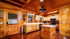 Interieur van een houten blokhut met een keuken, houten kasten, een roestvrijstalen koelkast en een houten deur met een kruismotief.