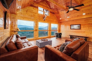 A cozy log cabin interior with leather sofas, a fireplace, and large windows offering mountain views.
