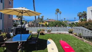 A patio with a table and chairs, a surfboard, and a view of the ocean. - WindanSea Ocean View (San Diego)
