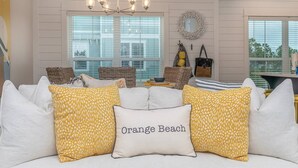 A living room with a white couch, yellow patterned pillows, a chandelier, wicker furniture, and a view through large windows.