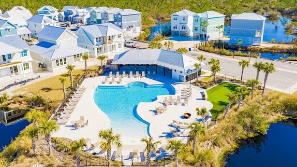 A resort with a large swimming pool surrounded by lounge chairs and palm trees, set against a backdrop of a forested area and a body of water.