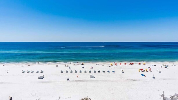 Ein Strand mit weißem Sand und klarem, blauem Wasser. Liegestühle und Personen befinden sich am Strand.