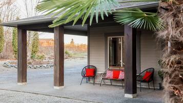 A covered patio with two wicker chairs and a table, a palm tree, and a building in the background.