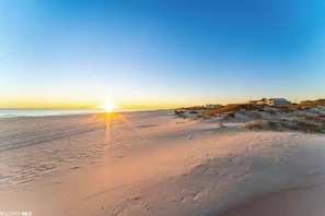 Una playa con un atardecer, dunas arenosas y un edificio al fondo.