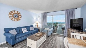 A living room with a blue sofa, a coffee table, a TV, and a balcony with a view of the ocean. - Plantation 6407-Beachfront Condo w/Heated Pool! (Gulf Shores)