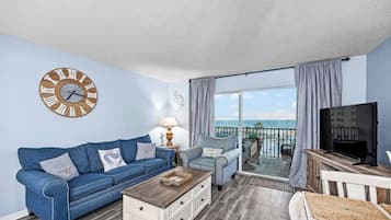 A living room with a blue sofa, a coffee table, a TV, and a balcony with a view of the ocean.