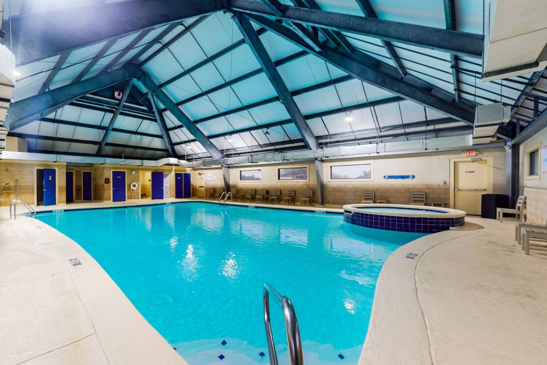 An indoor swimming pool with a high ceiling and skylights, surrounded by lounge areas and doors.