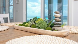 A wooden tray with a stack of stones and greenery on a wooden table, with a view of the ocean through large windows.