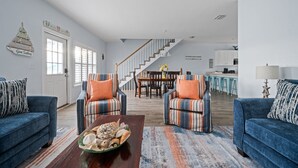 A spacious living room with a staircase leading to a kitchen, featuring blue sofas, striped armchairs, and a wooden coffee table with decorative items.