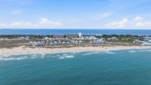 Vue aérienne d’une zone côtière avec des propriétés situées en bord de mer, une jetée et un grand bâtiment en arrière-plan.