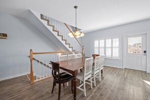 Un coin repas avec une table et des chaises en bois, un escalier avec des rampes blanches et une porte avec des volets blancs.