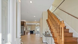 A spacious hallway with wooden flooring, a staircase with wooden balusters, and a modern kitchen in the background. - Spacious 3BR Home near Tysons and Metro | Private Yard (Falls Church)
