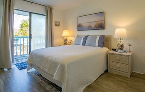 A bedroom with a large bed, two bedside tables with lamps, a painting on the wall, and a view through a sliding door to an outdoor area.