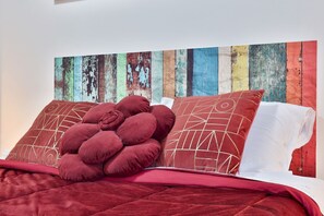 Ein Bett mit roter Decke und Kissen, eine Holzwand mit abblätternder Farbe und ein Kissen mit geometrischem Muster.