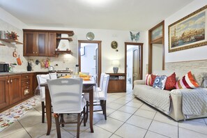 A modern kitchen with wooden cabinets, a dining table with chairs, a sofa with patterned cushions, and a painting of a cityscape.