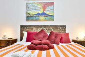 A bed with orange and white striped bedding, red pillows, and a painting of a volcano on the wall.