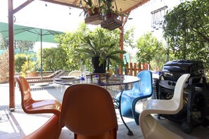 A covered outdoor seating area with a glass table, plastic chairs, and a barbecue grill.