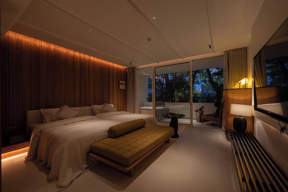 A modern bedroom with a large bed, a bench, a small table, and a view of the outdoors.