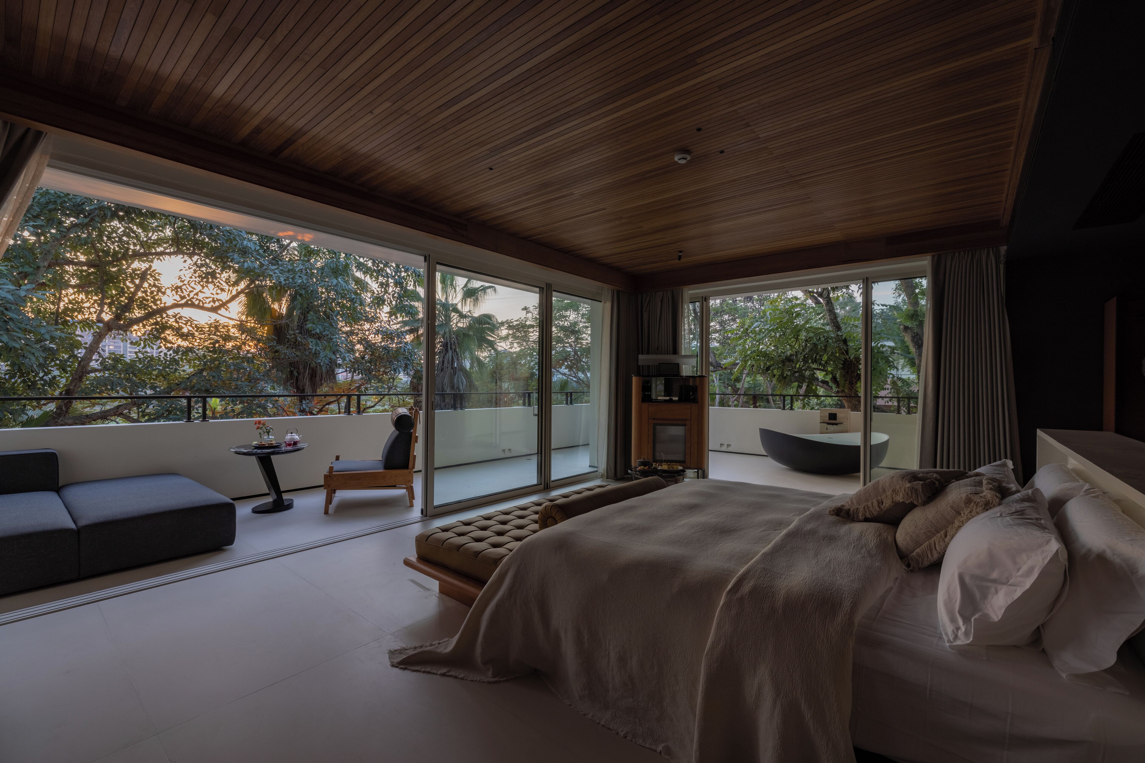 Een moderne slaapkamer met een groot bed, een sofa en een kleine tafel. Er is een balkon met uitzicht op bomen en een zwembad.
