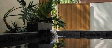 Un espace piscine paisible, agrémenté d’une clôture en bois, d’une végétation luxuriante et d’une petite cascade.