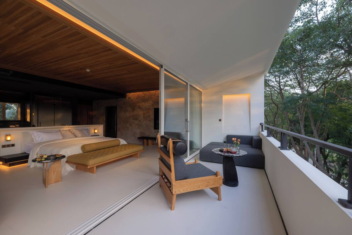 A modern bedroom with a bed, a wooden chair, a sofa, and a balcony with a railing.