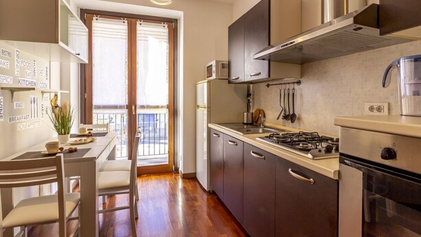 A modern kitchen with a gas stove, oven, and microwave, a dining area with a table and chairs, and a view through a glass door to an outdoor balcony.