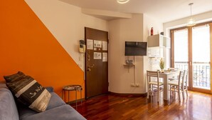 A living room with a sofa, a dining table with chairs, a TV mounted on the wall, and a door with a bulletin board.