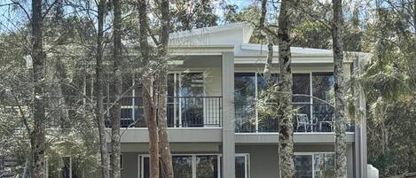 Una casa moderna de dos pisos con un balcón grande, rodeada de árboles y un césped bien cuidado.