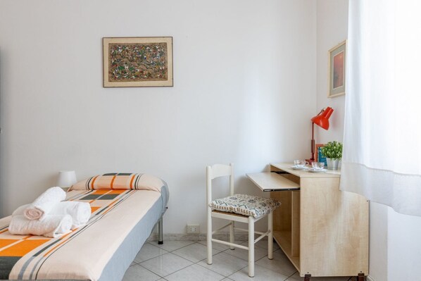 A bedroom with a bed, a desk, a chair, a small table, a lamp, and a framed picture on the wall.