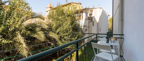 Un balcon avec une table et des chaises, entouré de verdure, et un bâtiment avec un toit en forme de dôme en arrière-plan.