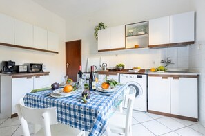 Une cuisine de taille réduite avec des meubles blancs, une table à manger recouverte d’une nappe à carreaux bleus et un lave-linge.