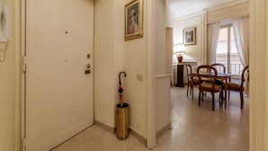 Un pasillo de hotel con una puerta cerrada, un cubo de basura, una zona de comedor con sillas y una mesa, y una ventana con cortinas.