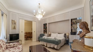 Una habitación bien iluminada con una cama, un sofá, un televisor, una araña y un busto sobre una mesa de mármol.