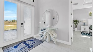 Un interior moderno con una mesa consola de vidrio, un espejo grande y vistas al exterior a través de una puerta.