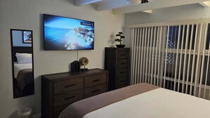 Een slaapkamer met een bed, een aan de muur bevestigde televisie, een plafondventilator, een dressoir en een raam met zonwering.