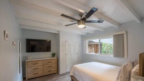 Ein Schlafzimmer mit einem Bett, einem Deckenventilator, einem Fernseher, einer hölzernen Kommode und einem Fenster mit Vorhängen.