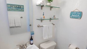 Ein Badezimmer mit einem weißen Waschbecken, einer Toilette und einem Spiegel. Oberhalb der Toilette befinden sich zwei Ablagen, eine mit einer Pflanze und die andere mit einem Handtuch. Ein Schild an der Wand zeigt die Aufschrift: I love you to the beach and back.