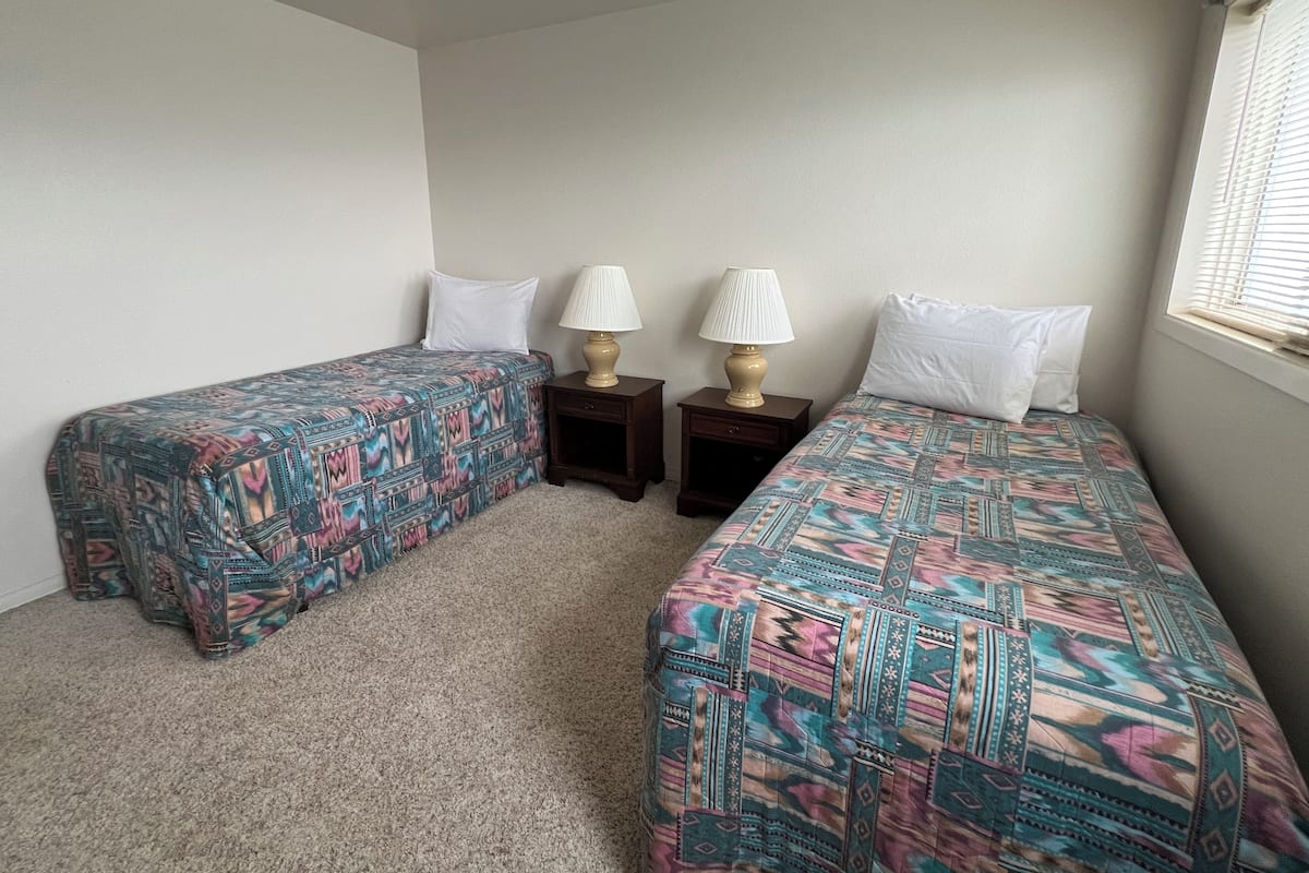 A room with two single beds, each with patterned bedding and white pillows. There are two bedside tables with lamps, and a window with blinds.