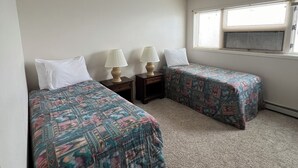 A small, clean room with two beds, each with a patterned bedspread and white pillows. There are bedside tables with lamps, a window with blinds, and a ceiling vent. - Adak Island Inn (Adak Island)